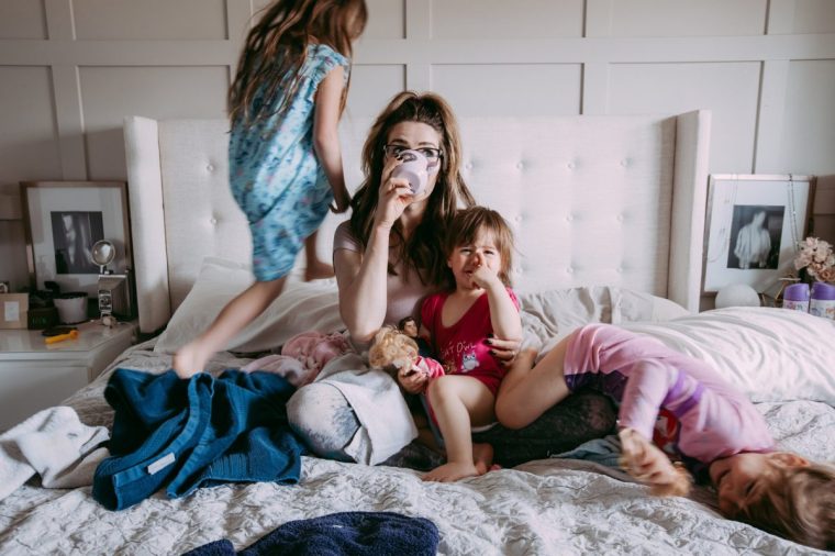 A picture of a young mother sipping a cup of coffee on a made-up bed while her three daughters play around her.