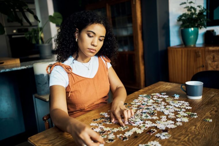 Woman working on a puzzle at the table in the living room of her modern comfortable home. Girl playing jigsaw puzzle challenge for mental health and relaxing while drinking cup of coffee in lounge at home