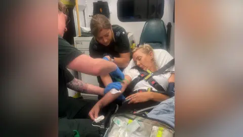 Michelle Swords of the family is assisted by paramedics. He lies unconscious, strapped to the ambulance bed. A man and a woman paramedic help him.