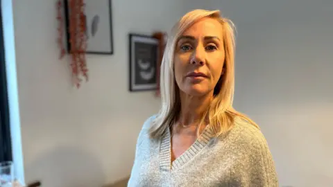 Michelle Talwar looks into the camera. She is in her house. Some pictures and decorations can be seen on the wall behind it.